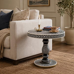 May include: A round side table featuring a black and white chevron pattern inlay across the top, pedestal base, and circular bottom. The table surface holds a brass candlestick with a white taper candle and a ceramic cup on a matching saucer.