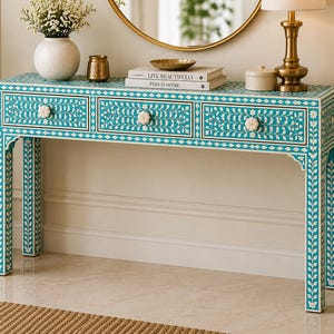 May include: A turquoise console table with three drawers, adorned with a floral pattern and white accents. The table features white flower-shaped drawer pulls and is placed against a white wall. A gold-toned mirror and lamp are visible in the background.