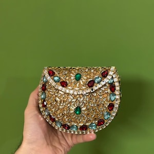 May include: A gold-toned, semi-circular clutch bag featuring an elaborate design of clear, red, green, and blue gemstones. The bag has a metallic finish and is set against a green backdrop.