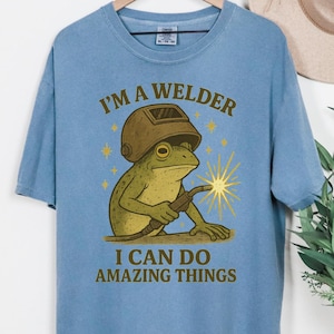 May include: Blue jean t-shirt featuring a frog welder graphic with the text "I'm a Welder I Can Do Amazing Things". The shirt is displayed on a wooden hanger. The frog is wearing a welding helmet and holding a torch.