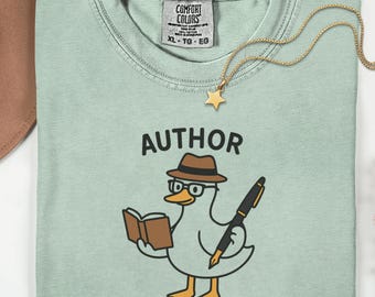 May include: A pale green t-shirt featuring the word "AUTHOR" above a cartoon duck wearing glasses and a hat, holding a book and a pen. A gold necklace with a star pendant is draped on the shirt. The shirt has a Comfort Colors tag.