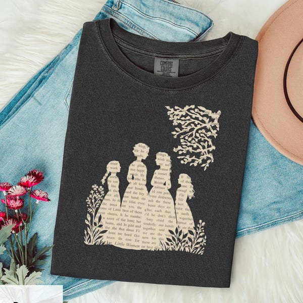 Puede incluir: Una camiseta gris carbón con un estampado gráfico de cuatro siluetas femeninas rellenas con texto del libro Mujercitas. El diseño incluye una rama de árbol sobre las figuras y elementos florales en la base del estampado.
