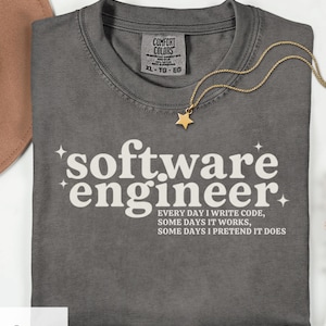 May include: A gray Comfort Colors t-shirt with the text "software engineer" and the phrase "EVERY DAY I WRITE CODE, SOME DAYS IT WORKS, SOME DAYS I PRETEND IT DOES." A gold star pendant necklace rests on the shirt.