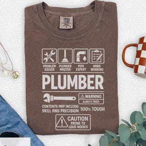 Pode incluir: Camiseta cor de café expresso com texto e gráficos brancos relacionados à canalização. A camisa inclui a palavra "PLUMBER" e frases como "Problem Solver" e "100% Tough". Uma caneca de café com um padrão xadrez marrom e branco é visível.