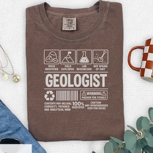 May include: A brown t-shirt with the word "GEOLOGIST" in white block letters. The shirt features icons and text including "Rock Identifier," "Field Explorer," and "Lab Researcher." The shirt also has the text "Warning: Passion for fossils."