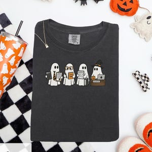 Può includere: Una t-shirt grigio antracite con una grafica di quattro fantasmi bianchi che svolgono compiti d'ufficio. Un fantasma indossa una cravatta e tiene una calcolatrice, un altro indossa occhiali e un libro, un terzo tiene delle fatture e il quarto indossa un cappello da strega a una scrivania.