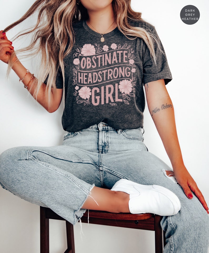 May include: A dark grey heather t-shirt with the text "Obstinate Headstrong Girl" in pink lettering surrounded by pink floral designs.