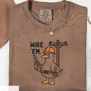 Puede incluir: Camiseta color expreso con un pato de dibujos animados que lleva un casco naranja y sostiene unos alicates. La camiseta presenta el texto "WIRE 'EM" y un collar con colgante de estrella dorada. Talla XL.