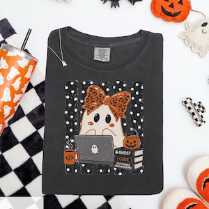 Op de afbeelding: Een antracietgrijs t-shirt met een afbeelding van een geest met een strik met luipaardprint, werkend op een laptop met een geeststicker. Het ontwerp bevat een mok met codesymbolen, een stapel boeken met de tekst Ghost Code Software Engineering en een pompoenemmer, alles tegen een achtergrond van zwart-witte stippenstrepen.