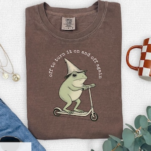 May include: Espresso-coloured t-shirt featuring a green frog in a wizard hat riding a scooter. The text "off to turn it on and off again" surrounds the frog. The shirt has a Comfort Colors label. A mug is visible.