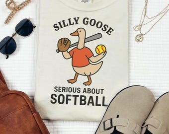 Puede incluir: Camiseta color marfil con un dibujo de un ganso con una camisa roja, sosteniendo un bate, un guante y una pelota de béisbol. El texto dice "Silly Goose Serious About Softball". La imagen también muestra gafas de sol, una pinza para el pelo, un collar, un bolso y sandalias.