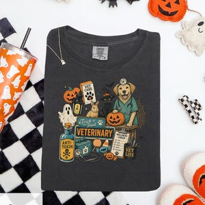 May include: A charcoal gray t-shirt featuring a graphic print of a golden retriever veterinarian, a black cat in a witch hat, a rabbit, a ghost, pumpkins, a medical kit, a stethoscope, and text that reads Trick or Treat Veterinary and Vet Life.