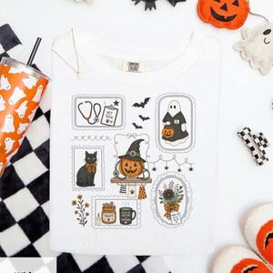 Puede incluir: Una camiseta blanca con un estampado gráfico de varias ilustraciones temáticas de Halloween dispuestas en marcos. El diseño incluye un estetoscopio, un portapapeles con la etiqueta Vet Tech, un fantasma, un gato negro, una calabaza y un ramo.