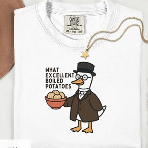 Może przedstawiać: Biała koszulka z grafiką kaczki w garniturze, kapeluszu i okularach, trzymającej miskę ziemniaków. Tekst brzmi "WHAT EXCELLENT BOILED POTATOES". Złoty naszyjnik z zawieszką w kształcie gwiazdy leży na koszulce.