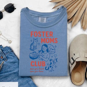 Pode incluir: Uma camiseta azul Comfort Colors com as palavras "FOSTER MOMS CLUB" em laranja, juntamente com ilustrações de gatos e uma mulher segurando um gato. O texto "open your heart, open your home" está abaixo.