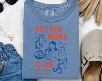 Puede incluir: Una camiseta azul Comfort Colors con las palabras "FOSTER MOMS CLUB" en naranja, junto con ilustraciones de gatos y una mujer sosteniendo un gato. El texto "open your heart, open your home" está debajo.