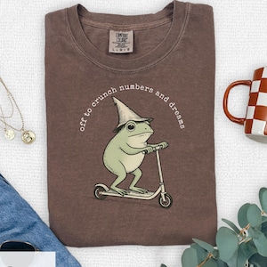 May include: Espresso-colored t-shirt with a whimsical graphic of a frog wearing a wizard hat and riding a scooter. The text "off to crunch numbers and dreams" is printed around the frog. A Comfort Colors tag is visible at the top.