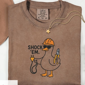 Puede incluir: Camiseta color expreso con un pato de dibujos animados que lleva un casco naranja y gafas de sol, sosteniendo un enchufe y unos alicates. El texto "SHOCK 'EM." está encima del pato. Un collar con colgante de estrella dorada está sobre la camiseta.