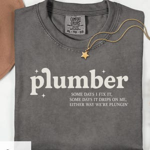 May include: A gray Comfort Colors t-shirt with the word "plumber" in white script. The shirt also features the text "SOME DAYS I FIX IT, SOME DAYS IT DRIPS ON ME, EITHER WAY WE'RE PLUNGIN'". A gold star pendant necklace rests on the shirt.