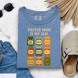 Op de afbeelding: Een blauw Comfort Colors T-shirt met de tekst "FOSTER MOM IS MY JAM" en illustraties van jampotten. Elke pot bevat een woord zoals "Guidance" of "Empathy". Het shirt wordt getoond met een spijkerbroek en accessoires.