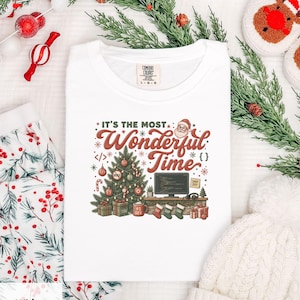 Può includere: Una t-shirt bianca con una stampa grafica che riporta il testo 'IT'S THE MOST Wonderful Time'. Il design include un albero di Natale decorato, un monitor di computer con del codice, un'icona di Babbo Natale, regali e calze appese.