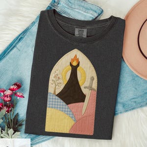 May include: Dark gray Comfort Colors t-shirt featuring a colorful, stitched design of a mountain with a flame, a sword, and a tree. The design is set within an arched frame. The shirt is folded on a white surface with blue jeans and a pink hat.