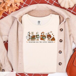Puede incluir: Una camiseta de cuello redondo color marfil con un gráfico de cuatro patos blancos vestidos como personal médico cuidando pavos pequeños. El texto debajo de la ilustración dice "TREATING ALL THE LITTLE TURKEYS" en una fuente de color marrón.
