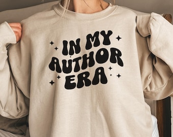 May include: A beige sweatshirt with the text "IN MY AUTHOR ERA" in black, retro-style lettering with stars on either side. The sweatshirt has a relaxed fit and is made of a soft, comfortable fabric.