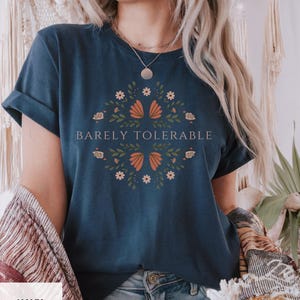 Puede incluir: Camiseta azul marino con mangas enrolladas. La camiseta presenta un diseño floral con detalles en naranja y blanco, y las palabras "Barely Tolerable" en el centro. La camiseta se combina con unos vaqueros azules.