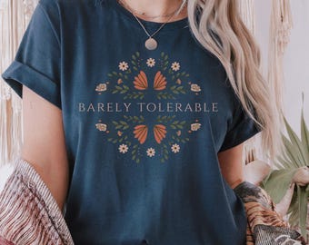 May include: Navy blue t-shirt with rolled sleeves. The shirt features a floral design with orange and white accents, and the words "Barely Tolerable" in the center. The shirt is paired with blue jeans.