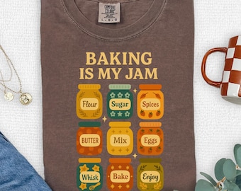 Puede incluir: Camiseta color espresso con el texto "BAKING IS MY JAM" e ilustraciones de ingredientes para hornear en frascos. Los frascos incluyen harina, azúcar, especias, mantequilla, mezcla, huevos, batidora, hornear y disfrutar.