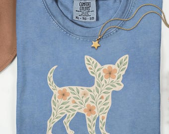 May include: A blue Comfort Colors t-shirt with a floral chihuahua design. The dog silhouette is filled with beige leaves and orange flowers. A gold star pendant necklace rests on the shirt. The shirt is labeled "Blue Jean".