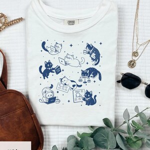 May include: A white t-shirt featuring a dark blue graphic print of various cats engaged in office and coding activities, including cats with laptops, a book labelled Clean Code, a scale, and a whiteboard with the text Agile.