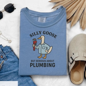 Puede incluir: Una camiseta Comfort Colors azul claro con un dibujo de un ganso sosteniendo una llave inglesa. El texto en la camiseta dice "SILLY GOOSE BUT SERIOUS ABOUT PLUMBING". La camiseta está doblada y se muestra sobre una superficie blanca.
