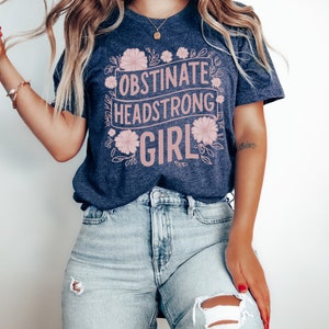 May include: A heather navy blue t-shirt with the text "Obstinate Headstrong Girl" in pink lettering surrounded by pink floral designs.
