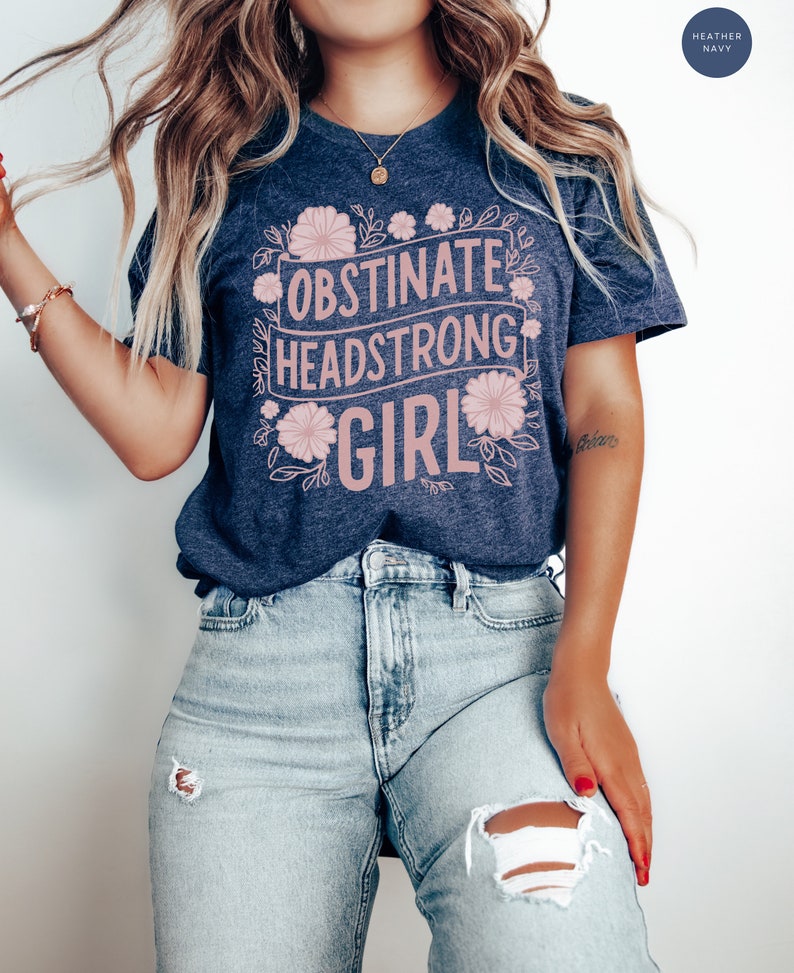 May include: A heather navy blue t-shirt with the text "Obstinate Headstrong Girl" in pink lettering surrounded by pink floral designs.