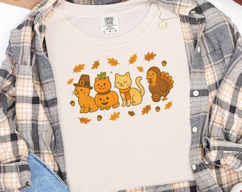 Könnte beinhalten: Elfenbeinfarbenes T-Shirt mit einem Thanksgiving-Motiv mit Cartoon-Katzen, die als Pilger, Kürbis und mit Schal verkleidet sind, zusammen mit einer Truthahn. Das Shirt ist mit einem karierten Hemd und Jeans überlagert. Ein Football und Herbstblätter sind ebenfalls sichtbar.