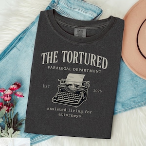 May include: A dark gray t-shirt with the text "THE TORTURED PARALEGAL DEPARTMENT" and a vintage typewriter illustration. The shirt also includes the text "EST 2026" and "assisted living for attorneys".
