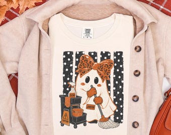 May include: An ivory t-shirt featuring a graphic of a ghost wearing a leopard print bow, holding a mop, and standing next to a cleaning cart. The ghost has a name tag that reads JANITOR. The background of the graphic has black and white polka dots.