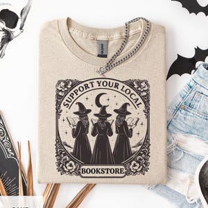 Puede incluir: Camiseta color arena con un gráfico que dice "Support Your Local Bookstore". El diseño presenta tres brujas leyendo libros bajo una luna creciente y estrellas. Un collar de cadena plateada descansa sobre la camisa.