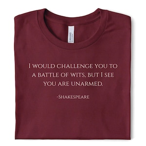 Puede incluir: Una camiseta de cuello redondo color granate con el texto impreso en blanco: I WOULD CHALLENGE YOU TO A BATTLE OF WITS, BUT I SEE YOU ARE UNARMED. -SHAKESPEARE. La camiseta tiene una etiqueta interior blanca en el cuello con la marca Canvas.