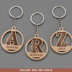 Puede incluir: Tres llaveros circulares de madera con las letras A, R y Z. Cada letra está grabada con un patrón floral y acompañada de un nombre: ADAM, RAVEN y ZET. Los llaveros incluyen una anilla de metal plateado sujeta por una cadena corta.