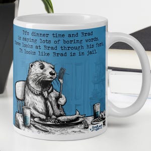 May include: White ceramic mug with a blue background and a cartoon illustration of an otter eating a fish. The text on the mug reads: "It's dinner time and Brad is saying lots of boring words. Steve looks at Brad through his fork. It looks like Brad is in jail."