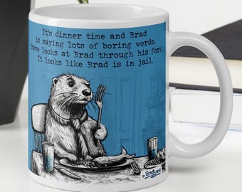 May include: White ceramic mug with a blue background and a cartoon illustration of an otter eating a fish. The text on the mug reads: "It's dinner time and Brad is saying lots of boring words. Steve looks at Brad through his fork. It looks like Brad is in jail."