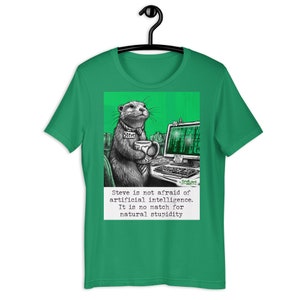 Puede incluir: Una camiseta verde con una ilustración de dibujos animados de una nutria con una etiqueta de nombre que dice "Steve". La nutria está sentada en un escritorio con una computadora y una taza de café. El texto debajo de la imagen dice "Steve no le teme a la inteligencia artificial. No es rival para la estupidez natural."