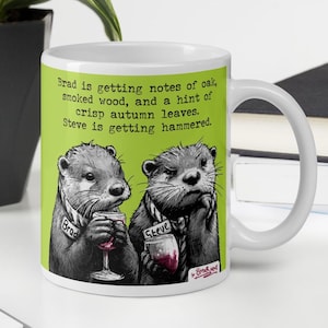 May include: White ceramic mug with a humorous design. It depicts two otters, one labeled "Brad" enjoying wine, the other, "Steve," looking unimpressed. The mug's text reads, "Brad is getting notes of oak... Steve is getting hammered."