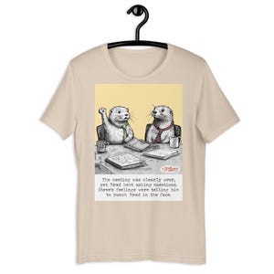 Op de afbeelding: Een beige t-shirt met een cartoonillustratie van twee otters in een vergadering. De otter links steekt zijn poot op en de otter rechts zit met zijn poten op een tafel. De tekst onder de afbeelding luidt: "De vergadering was duidelijk voorbij, maar Brad bleef vragen stellen. Steves gevoelens zeiden hem dat hij Brad in zijn gezicht moest slaan."