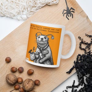 May include: White ceramic mug featuring a cartoon illustration of an otter wearing a scarf and holding a bottle and a mug. The otter has a name tag that says "Steve". The background is orange and the text on the mug reads "Steve has finally worked out what he'll be for Halloween: Intoxicated."