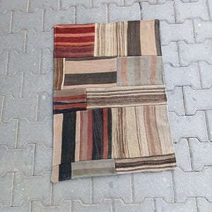 Könnte beinhalten: Ein rechteckiger Patchwork-Teppich mit einer Vielzahl von Farben und Mustern. Der Teppich hat Abschnitte mit roten, braunen, beigen und schwarzen Streifen und unifarbenen Farben. Der Teppich besteht aus einem gewebten Material.