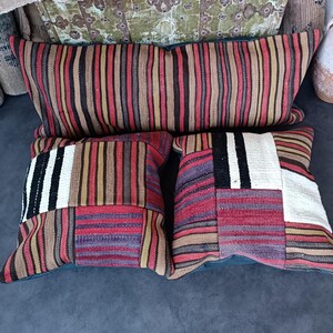 May include: Three decorative throw pillows with a striped and patchwork design in shades of red, black, white, brown, and beige. The pillows are arranged on a dark gray surface.